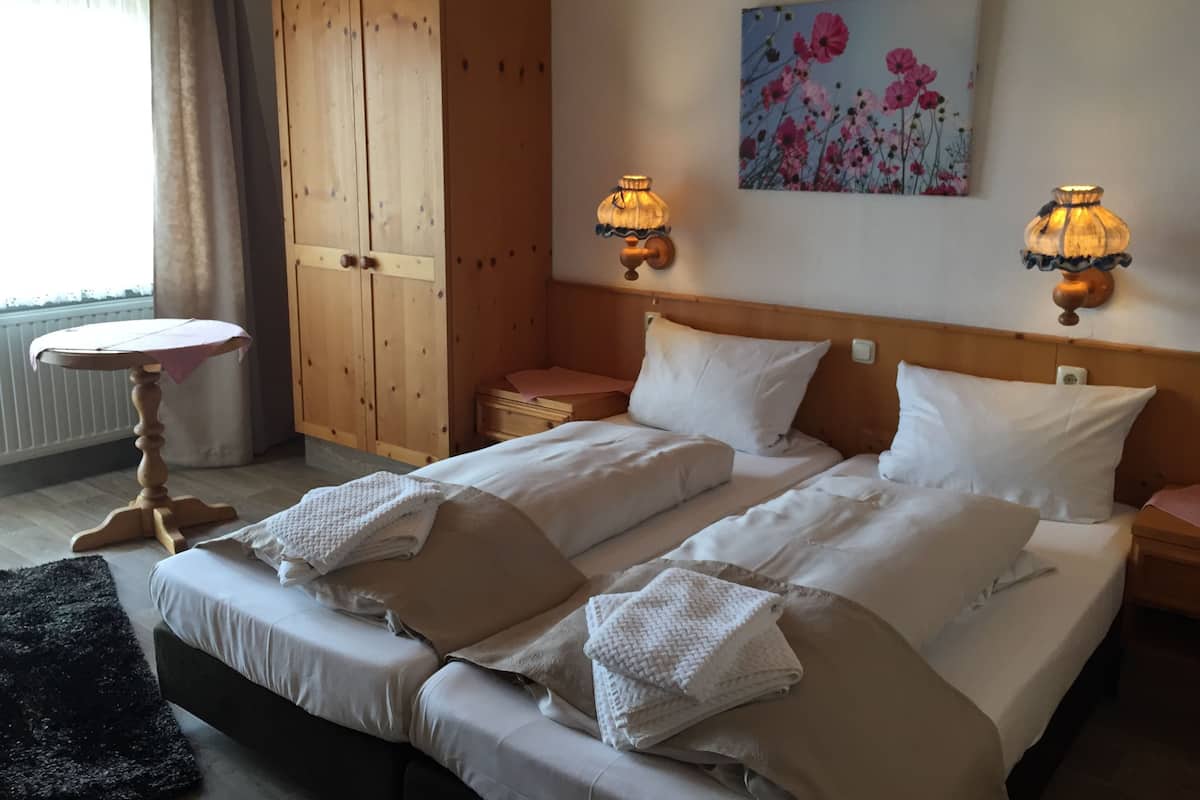 A hotel room with two beds, a wooden wardrobe, a small table, and two wall-mounted lamps.