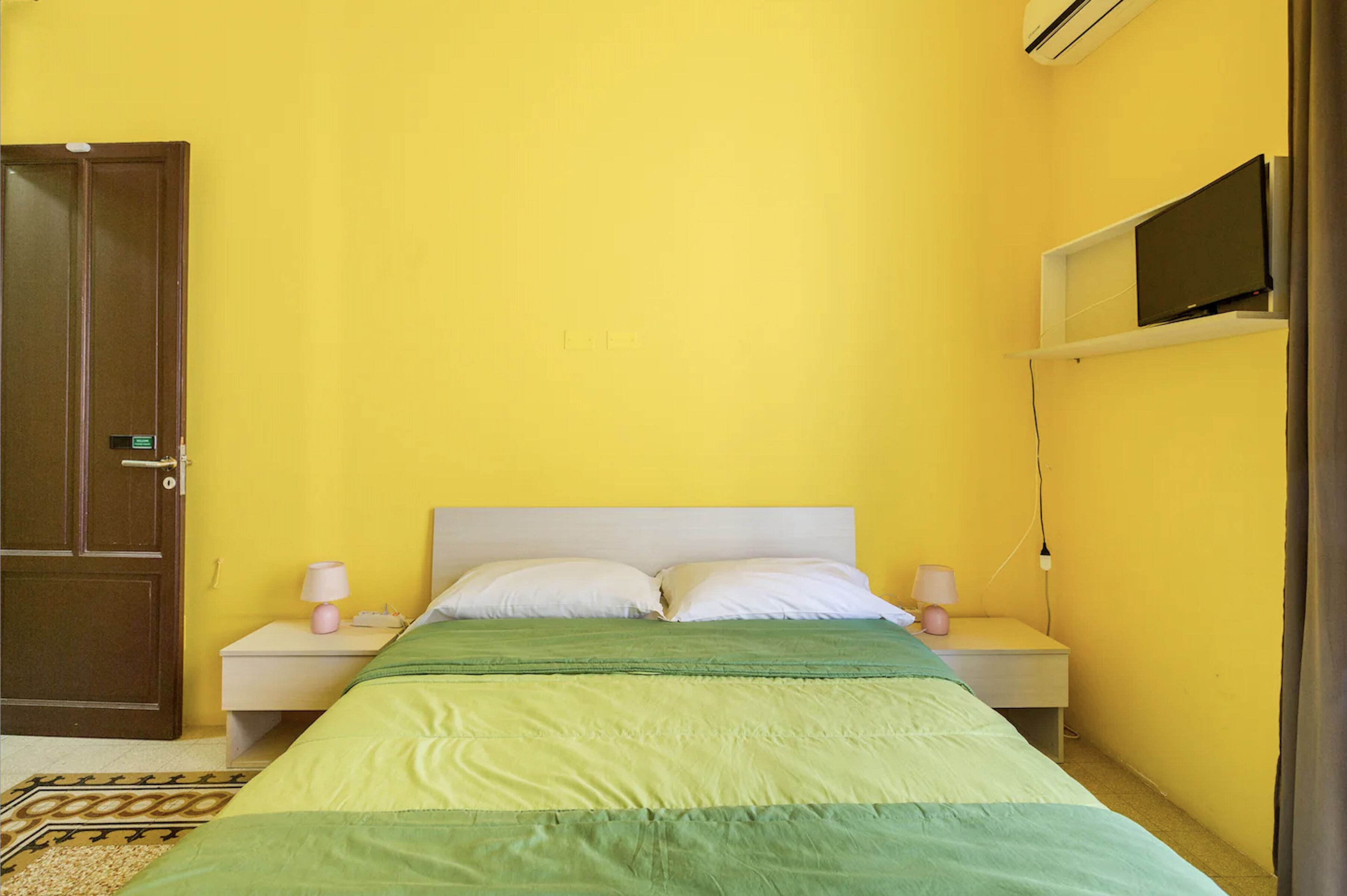 Une petite chambre avec un lit simple, un mur jaune, une porte en bois, une table de chevet blanche avec une lampe et un téléviseur fixé au-dessus du lit.