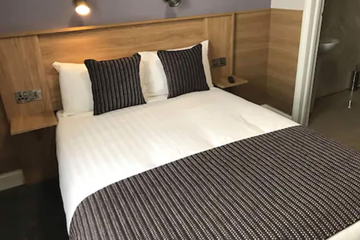 Habitación de hotel con cabecera de madera, cama con ropa de cama blanca y a rayas, mesita de noche pequeña y lámpara fijada a la pared.