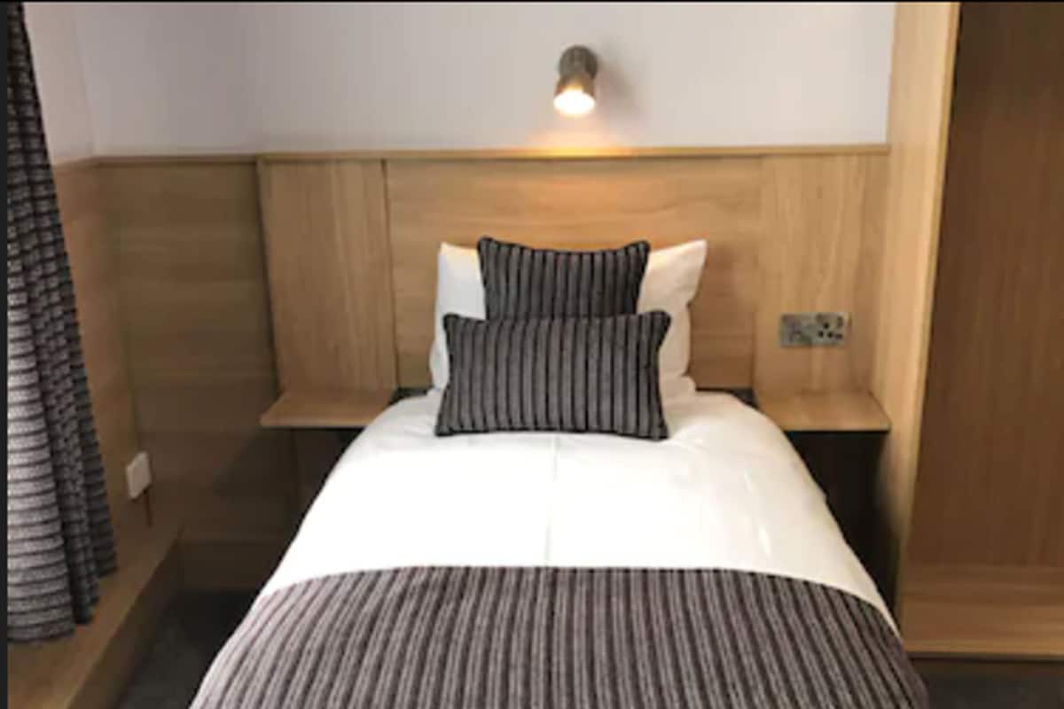 Una cama bien hecha con colcha a rayas y almohadas, una mesita de noche con lámpara y un cabecero de madera.