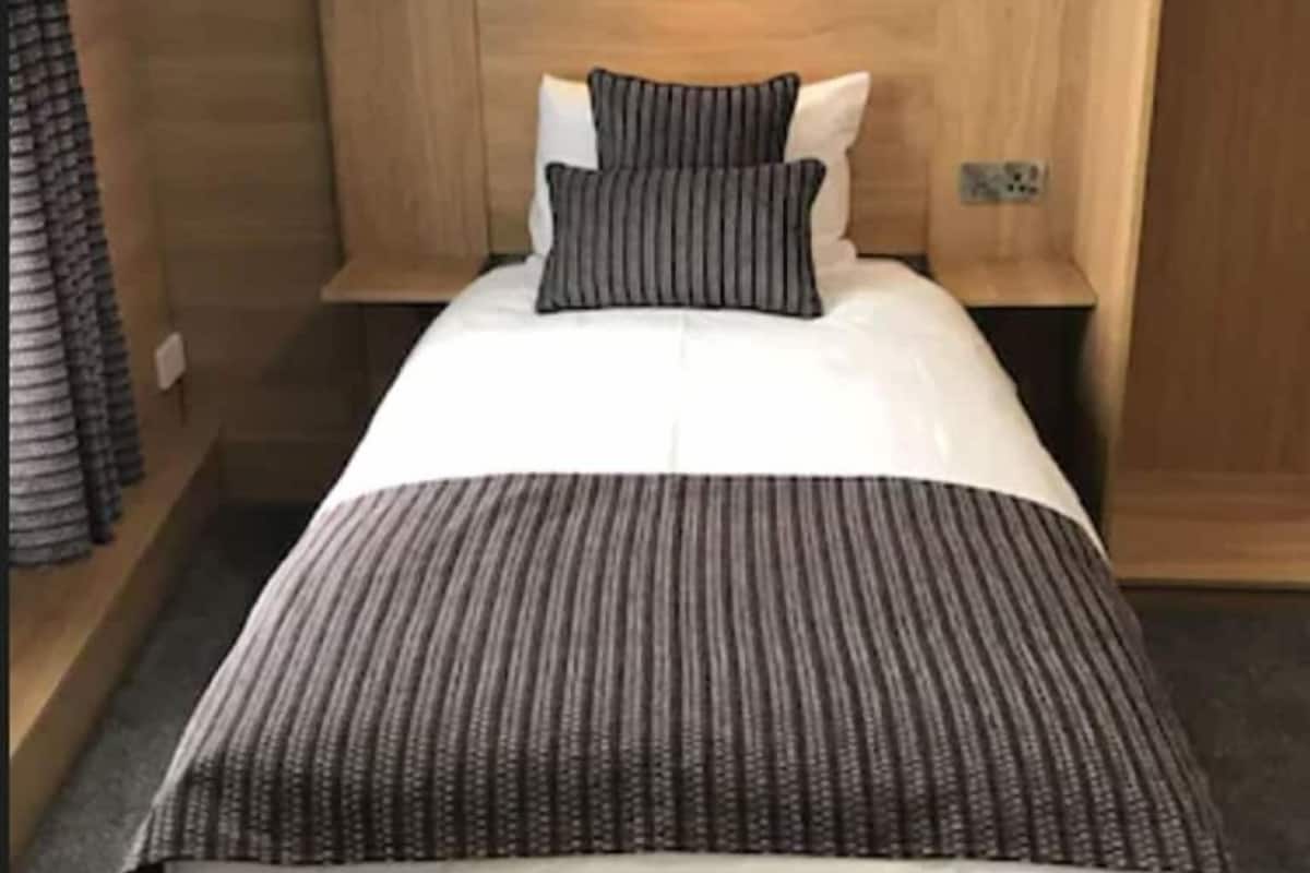 Una cama individual con una colcha a rayas, dos almohadas, una mesita de noche y una lámpara fijada a la pared.