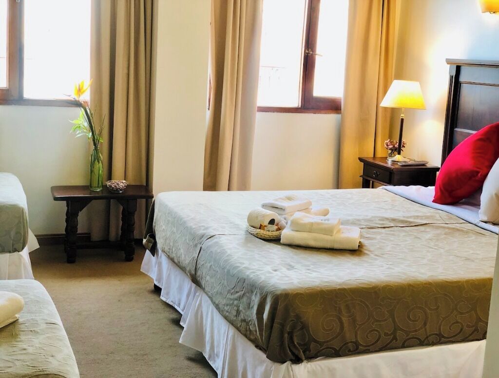 A hotel room with a bed, a bedside table with a lamp, a vase with flowers, and a window with curtains.