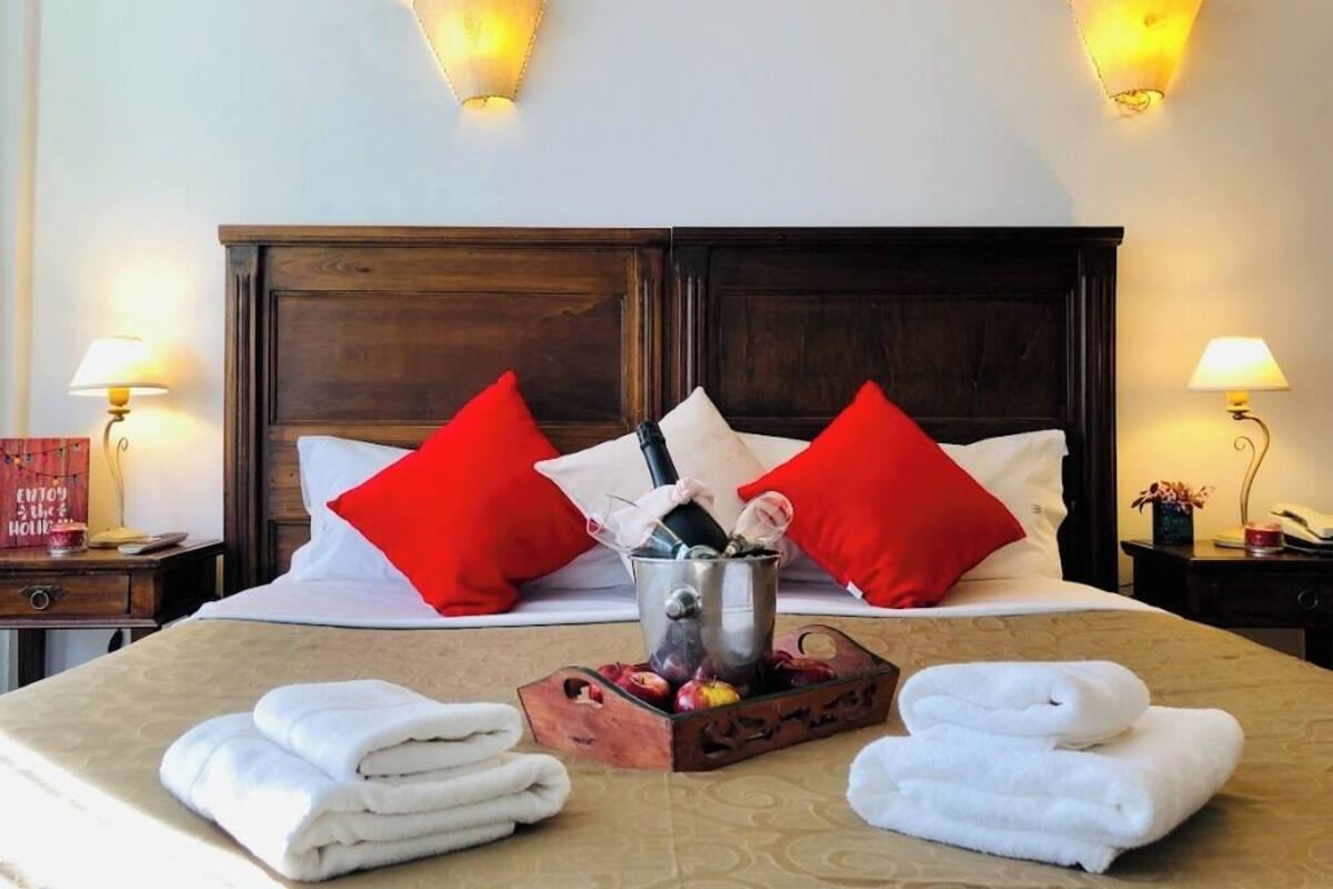 A hotel room with a bed, two red pillows, a bedside table with a lamp, a nightstand with a lamp, and a tray with towels and a bottle.
