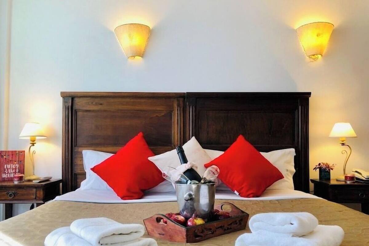 A hotel room with a bed, two red pillows, a bedside table with a lamp, a nightstand with a lamp, and a tray with towels and fruit.