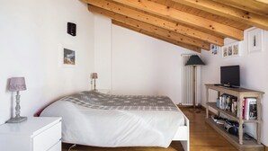 A bedroom with a wooden ceiling, a bed with a patterned blanket, a bedside table with a lamp, a floor lamp, a TV on a wooden stand, and a bookshelf. - Cozy attic among the rooftops of the city (Bologna)