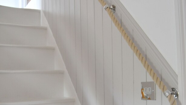 Un escalier en bois peint en blanc, doté d’une rampe en corde et d’un interrupteur sur le mur.