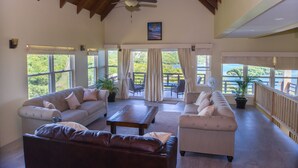 A spacious living area with two sofas, a coffee table, and a ceiling fan. Large windows provide views of the outdoors.