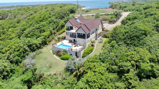 A two-story house with a swimming pool, surrounded by lush greenery and overlooking a coastal area.