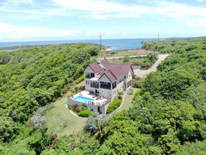 Une maison à deux étages avec une piscine, entourée d’une végétation luxuriante et offrant une vue sur la côte.