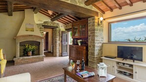 Un salon rustique avec des poutres apparentes, une cheminée en pierre, un meuble en bois et un téléviseur posé sur un meuble blanc.
