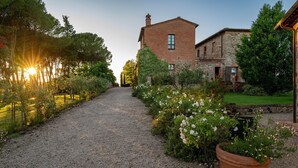 En grussti fører hen til en landlig bygning med murstensfacade og stenudsmykning, omgivet af frodig vegetation og blomstrende planter.