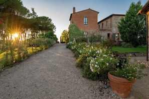 Un chemin de gravier mène à un bâtiment rustique à la façade en briques et aux détails en pierre, entouré d’une végétation luxuriante et de plantes en fleurs.