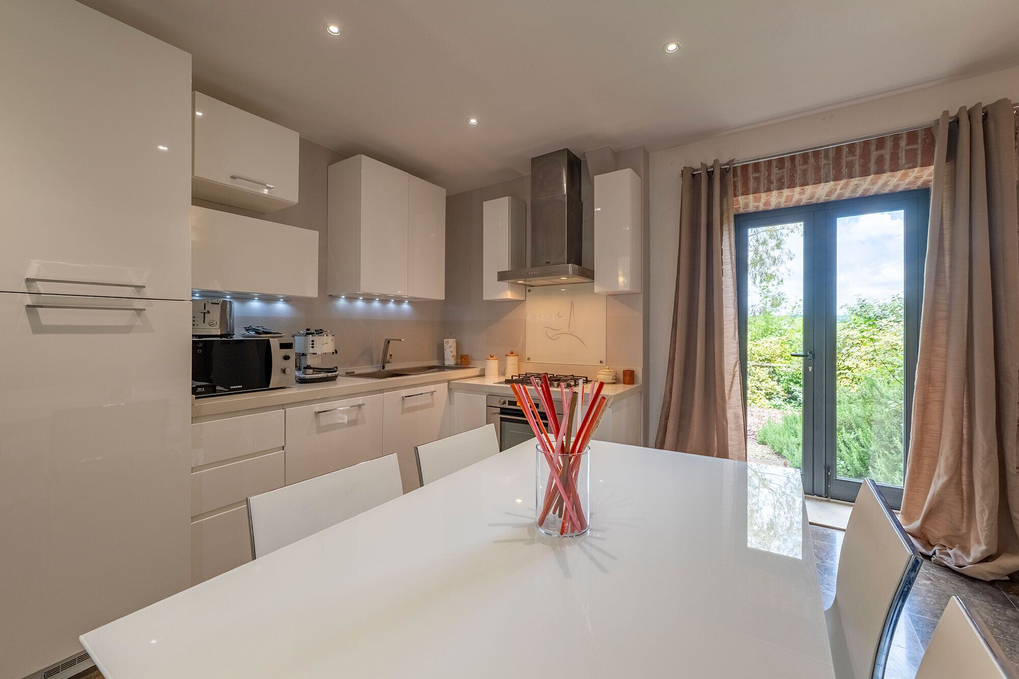 Una cocina moderna con armarios blancos, una mesa de comedor y vistas a la vegetación a través de un ventanal grande.