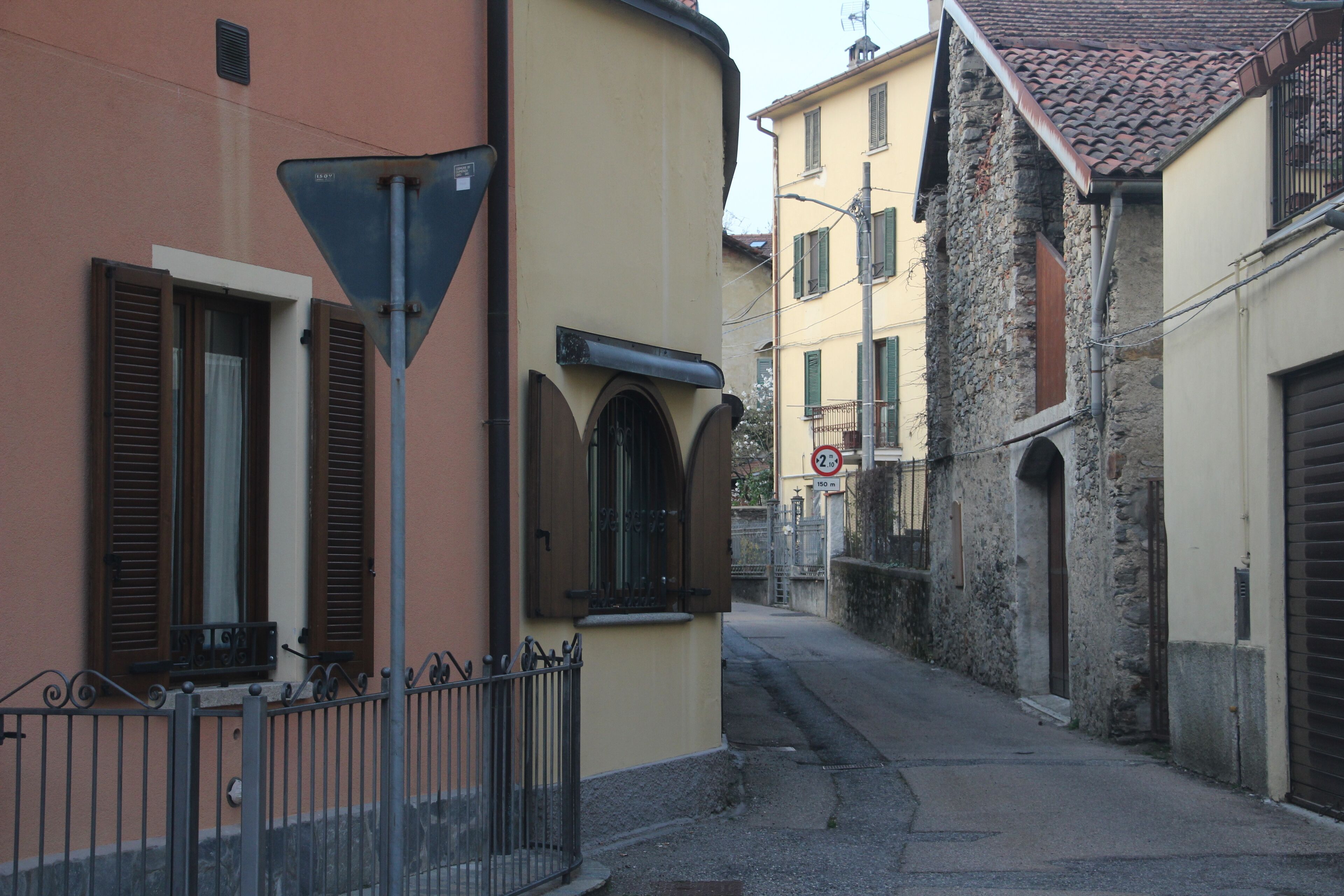 Una calle estrecha flanqueada por edificios, con fachadas de piedra y enlucido, una cerca metálica y un cartel de prohibido estacionar.