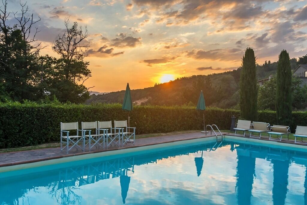 A swimming pool with lounge chairs and umbrellas, surrounded by greenery and a sunset view.