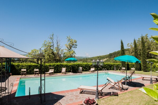 A swimming pool with lounge chairs and umbrellas, surrounded by greenery and a clear sky.