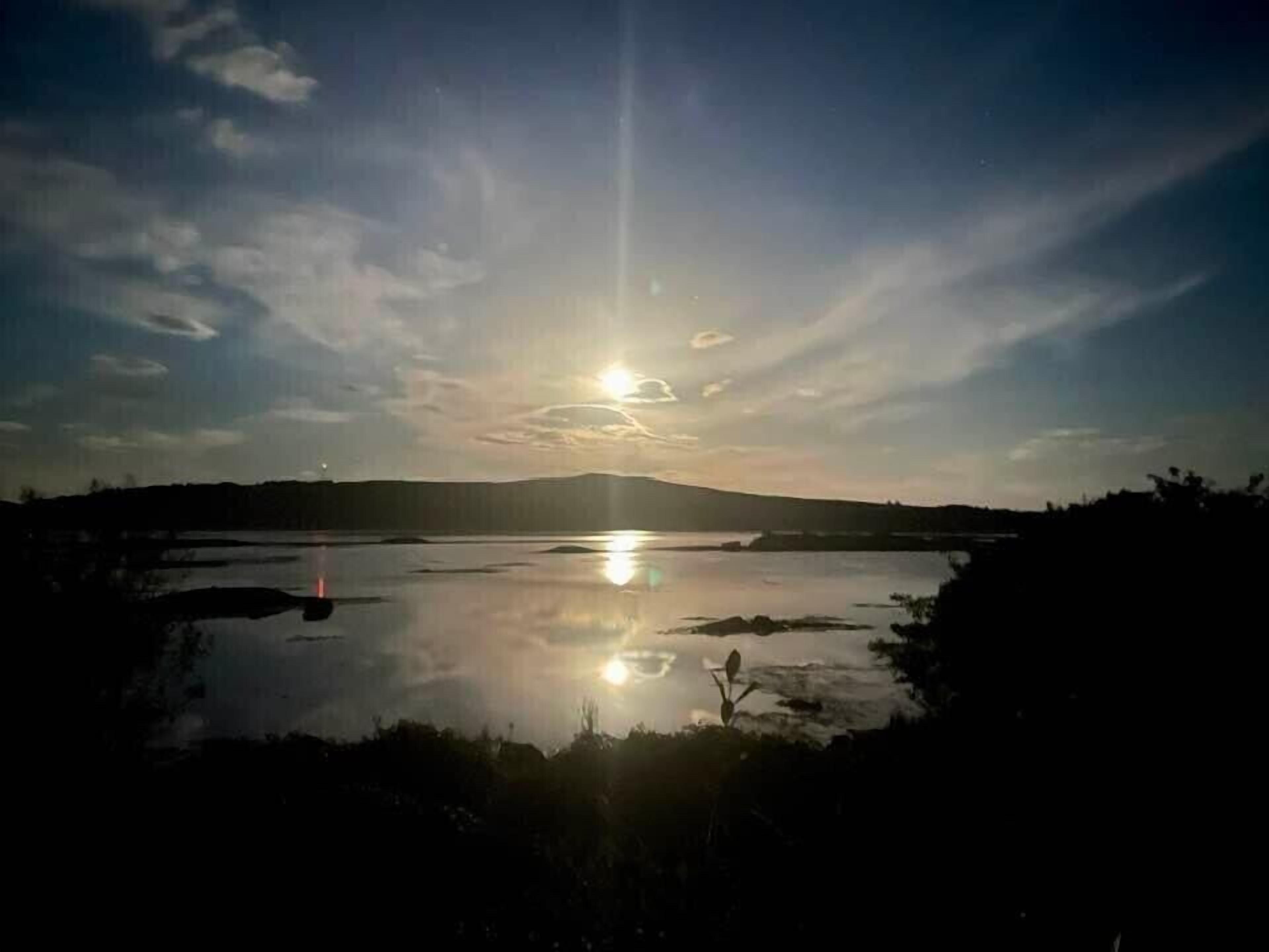 A calm lake with the sun reflecting on the water, surrounded by hills and sparse vegetation.