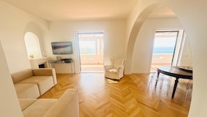 A spacious living area with a wooden herringbone floor, a beige sofa, a white armchair, and a dining table with chairs. There is a fireplace with a TV mounted above it, and a large window offering a view of the sea. - Beautiful panoramic villa in a private park with descent to the sea (Massa Lubrense)