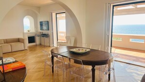 A spacious living area with a wooden floor, a round dining table with clear chairs, a sofa, and a fireplace. Large windows offer a view of the sea. - Beautiful panoramic villa in a private park with descent to the sea (Massa Lubrense)