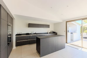 A modern kitchen with dark cabinetry, a central island, and a sliding glass door leading to an outdoor area. - 5* villa with private pool on prestigious estate (Villeneuve-Loubet)