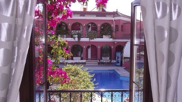 Une vue d’une piscine avec, en arrière-plan, un bâtiment rose, entouré de plantes et de fleurs en pot.