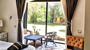 Un dormitorio con una cama, una silla, una mesita y vistas a un jardín a través de un ventanal.