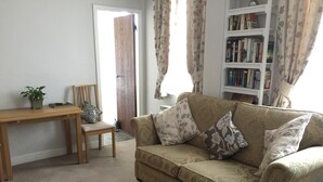 Ein Wohnzimmer mit einem beigen Sofa, einem Holztisch, einem Sessel, einem Bücherregal und einer Tür zu einem anderen Raum.