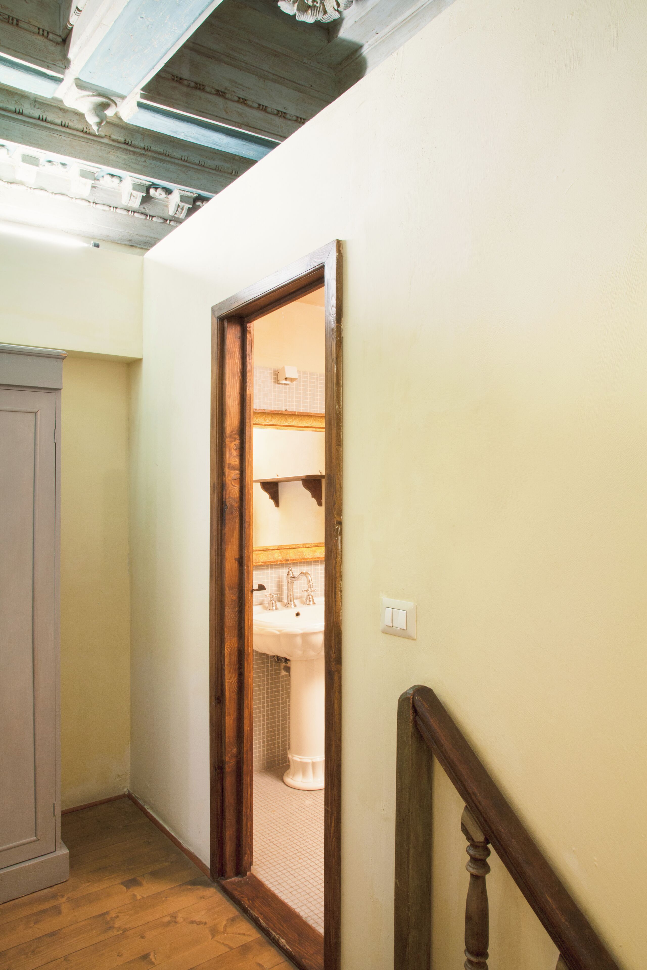 Un couloir étroit avec une porte en bois donnant sur une salle de bain équipée d’un lavabo et d’un miroir.