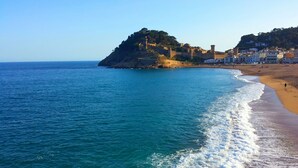 Un pueblo costero con un castillo imponente en la cima de una colina, una playa de arena y un mar azul cristalino.