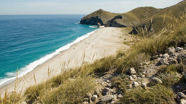 Una platja amb aigües turqueses cristal·lines, sorra daurada i turons rocosos al fons.
