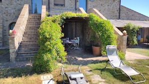 Un edificio de piedra con una escalera, un espacio al aire libre cubierto con una mesa y sillas, y un jardín con una silla y una jardinera.