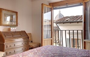 A bedroom with a bed, dresser, chair, and a balcony with a view of buildings.