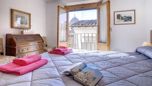 Een slaapkamer met een bed, een houten dressoir, een raam met uitzicht op een gebouw en een lijst met een afbeelding aan de muur.