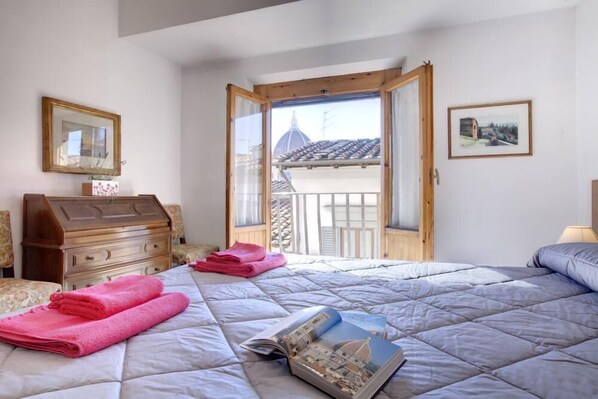 A bedroom with a bed, a wooden dresser, a window with a view of a building, and a framed picture on the wall.