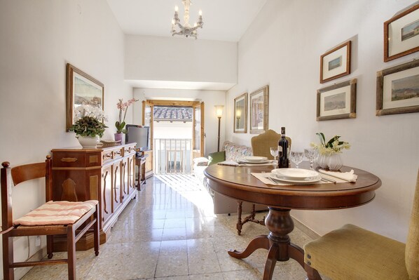 A dining area with a round table set for four, flanked by chairs. A wooden cabinet with drawers is on the left, and a sofa is in the background. The room has a high ceiling with a chandelier.
