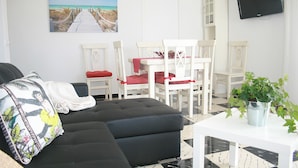 Uma sala de estar com sofá, uma mesa de jantar com cadeiras, uma televisão fixada na parede e um quadro de uma praia.