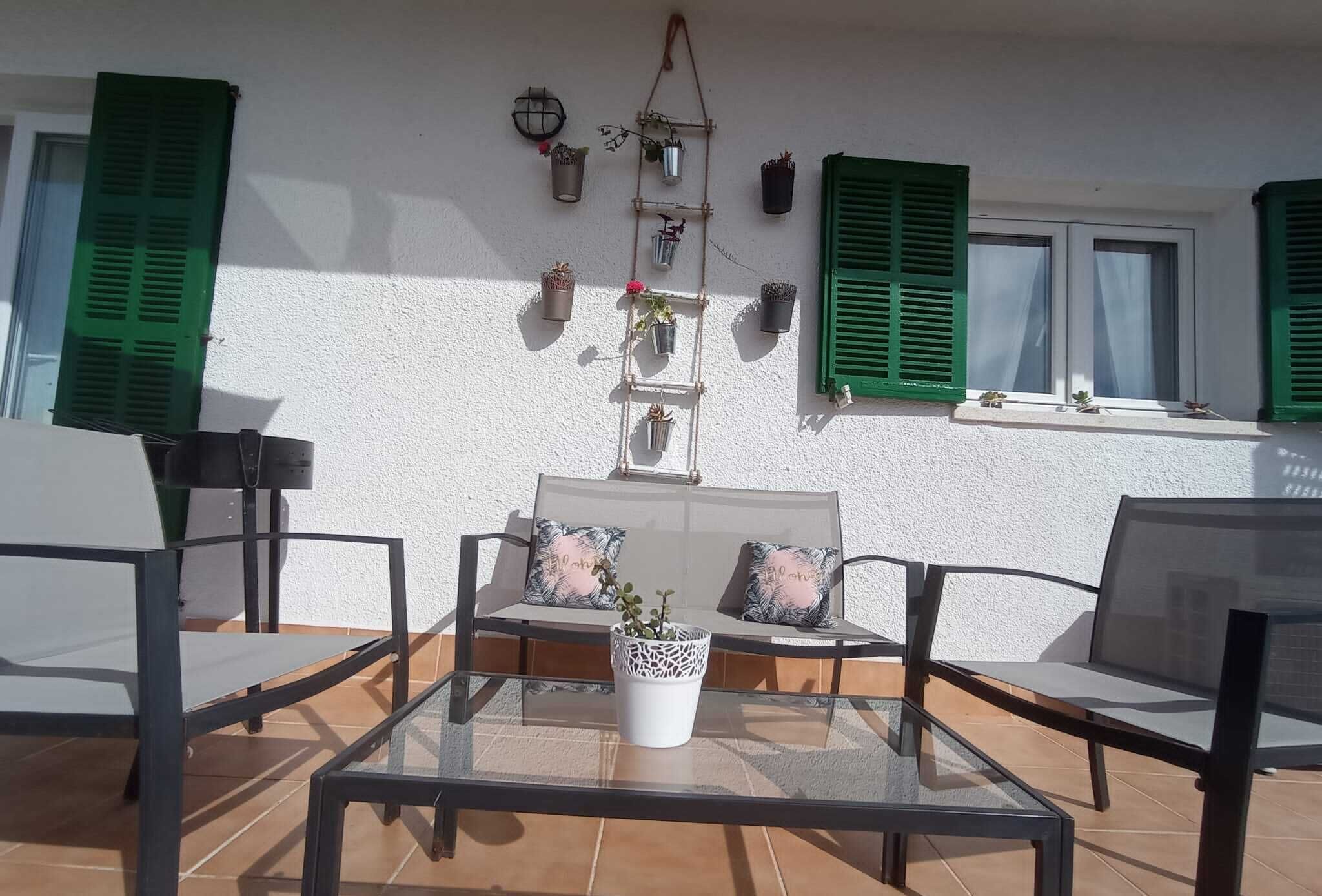 Une terrasse ensoleillée avec une table en verre, deux fauteuils rembourrés, un canapé agrémenté de coussins décoratifs et une étagère à plantes fixée au mur.