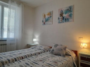 Un dormitorio con dos camas, una ventana con cortinas translúcidas, una mesita de noche con lámpara y dos cuadros colgados en la pared.