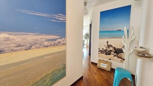 Una habitación con un ventanal grande, suelo de madera, un taburete azul y una estantería de pared con objetos decorativos.