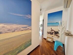 Una habitación con un ventanal grande, suelo de madera, un taburete azul y una estantería de pared con objetos decorativos.