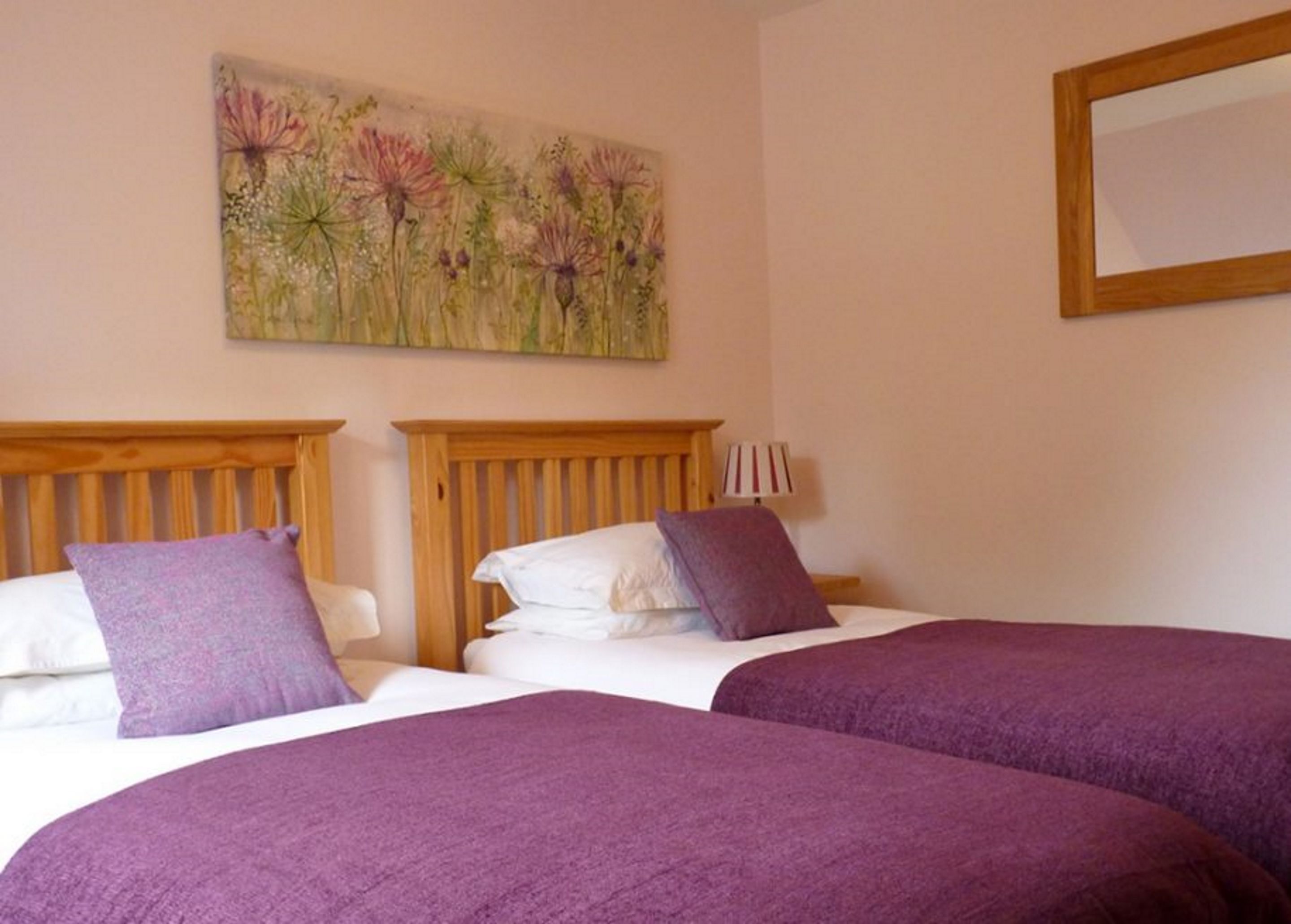 A hotel room with two beds, each with a wooden headboard, white linens, and purple bedding. A painting of flowers hangs on the wall above the beds.