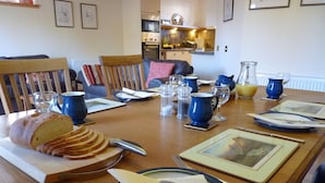 Una mesa de comedor dispuesta con pan, platos, cubiertos y vasos, con una cocina al fondo.