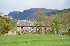 A stone house with a red door and chimney, surrounded by a grassy field and trees, with mountains in the background. - Traditional Family Friendly Farmhouse cottage - sleeps 8 (Santon Bridge, Cumbria)