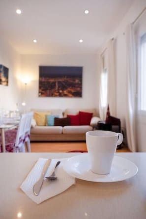 A dining table with a white cup and saucer, a knife, and a napkin. In the background, there is a sofa with colorful cushions and a cityscape painting on the wall.