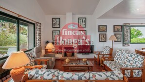 A spacious living room with a sofa set, a coffee table, and a television. There are framed pictures on the walls and a window with curtains.