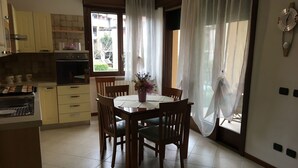 A dining area with a wooden table and chairs, a vase with flowers, and a view through a window with sheer curtains. - Gemma - apartment holiday home (Desenzano del Garda)