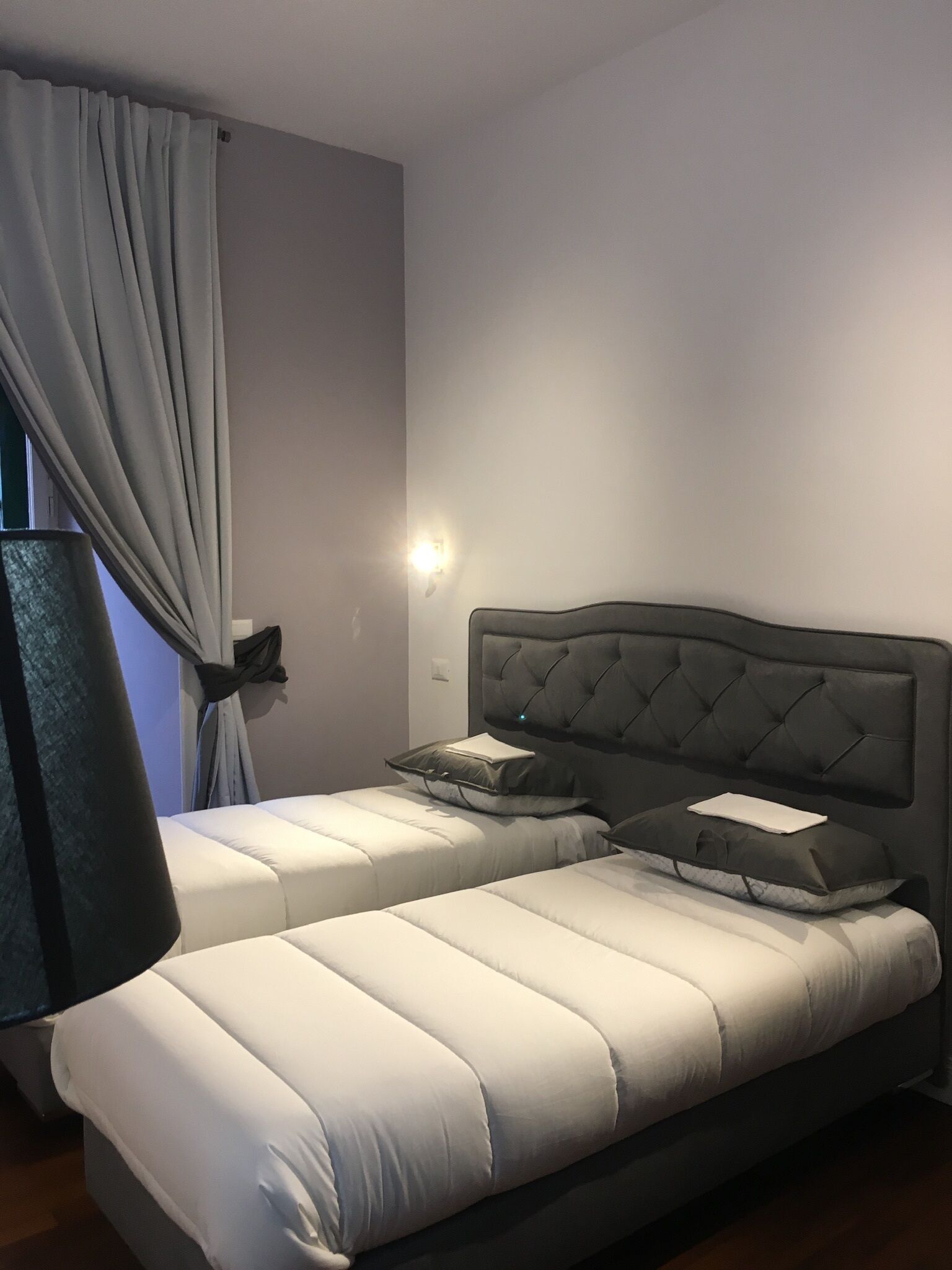 A bedroom with two beds, a large window with curtains, and a wall-mounted light.