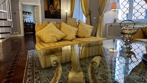 A room with a sofa, a glass coffee table, a patterned rug, and a view through a window with curtains.