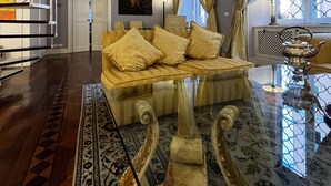 A spacious living room with a glass coffee table, a sofa with patterned cushions, a staircase, and a large window with curtains.