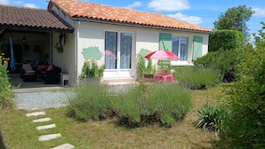 Une maison avec un toit en tuiles, un patio couvert avec des meubles d’extérieur et un jardin où poussent différentes espèces de plantes.